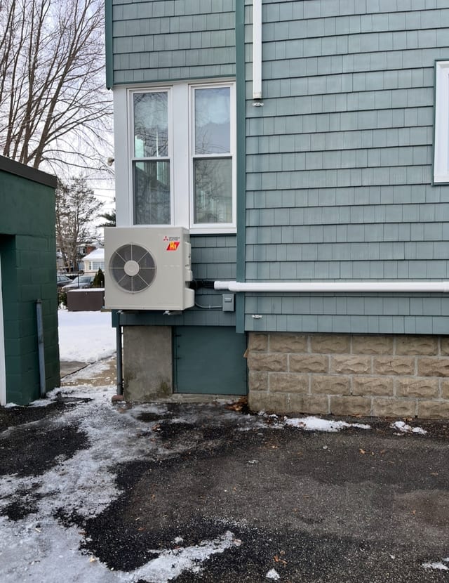 Mini split outdoor unit mounted on the foundation of a blue shingled house